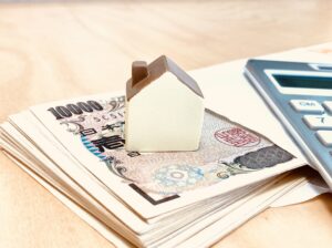 一万円札の束の上に置かれた小さな家の模型と電卓。不動産経営や不動産投資の基礎知識をイメージした写真