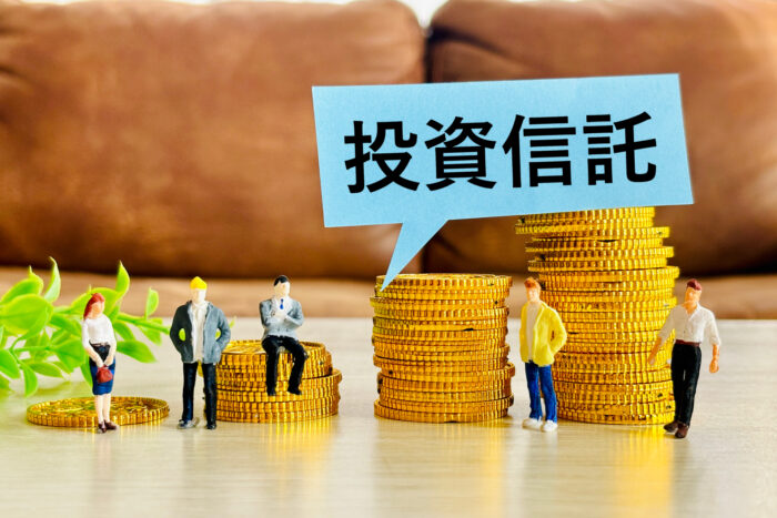 「投資信託」の吹き出しと硬貨の山の前に立つ人形のイメージ。NISAで不動産投資信託を組み込む割合を考える