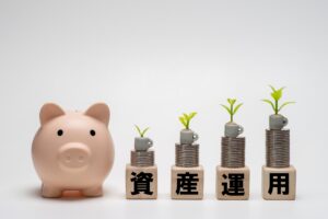 NISAの不動産投資信託で資産運用を始めるイメージ。貯金箱の横で、積み上げた硬貨から芽が育つ「資産運用」の文字ブロック